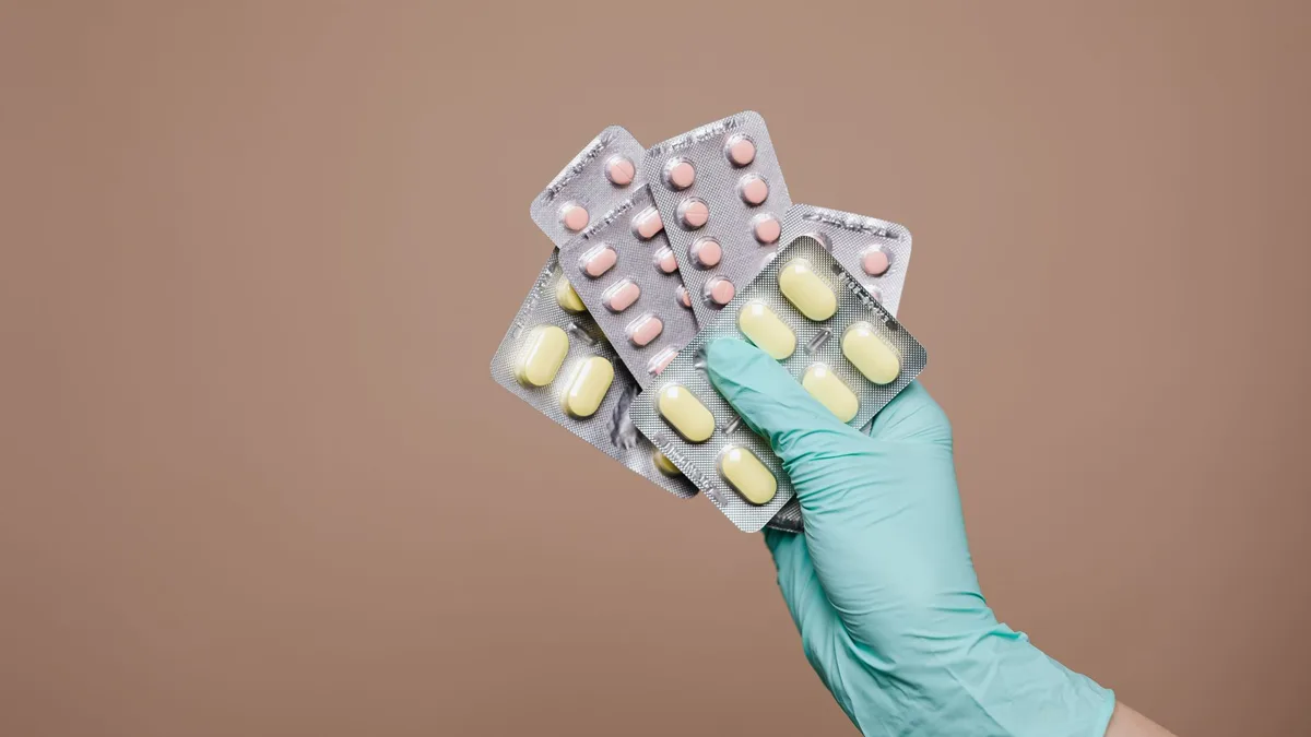Close-up of a gloved hand holding multiple blister packs of tablets against a neutral background.
