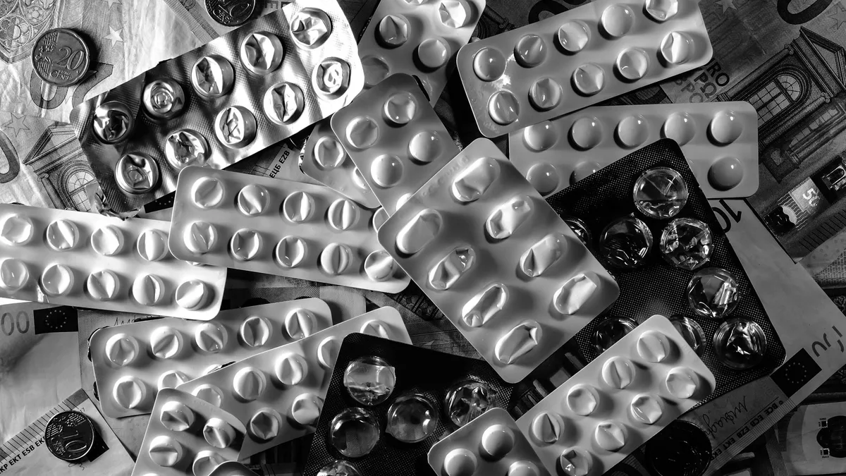 Black and white image of pill blister packs scattered on euro bills and coins.