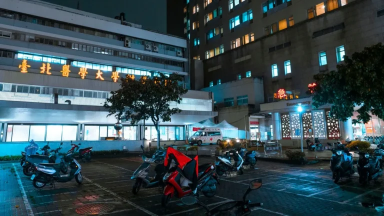Night view of a hospital with scooters parked outside, illuminated by bright city lights.