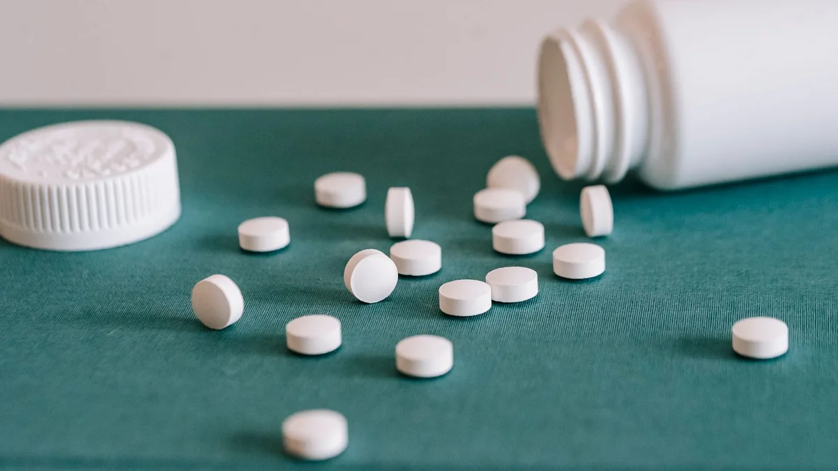 White medical pills spilled out of white plastic jar on green fabric surface against white wall